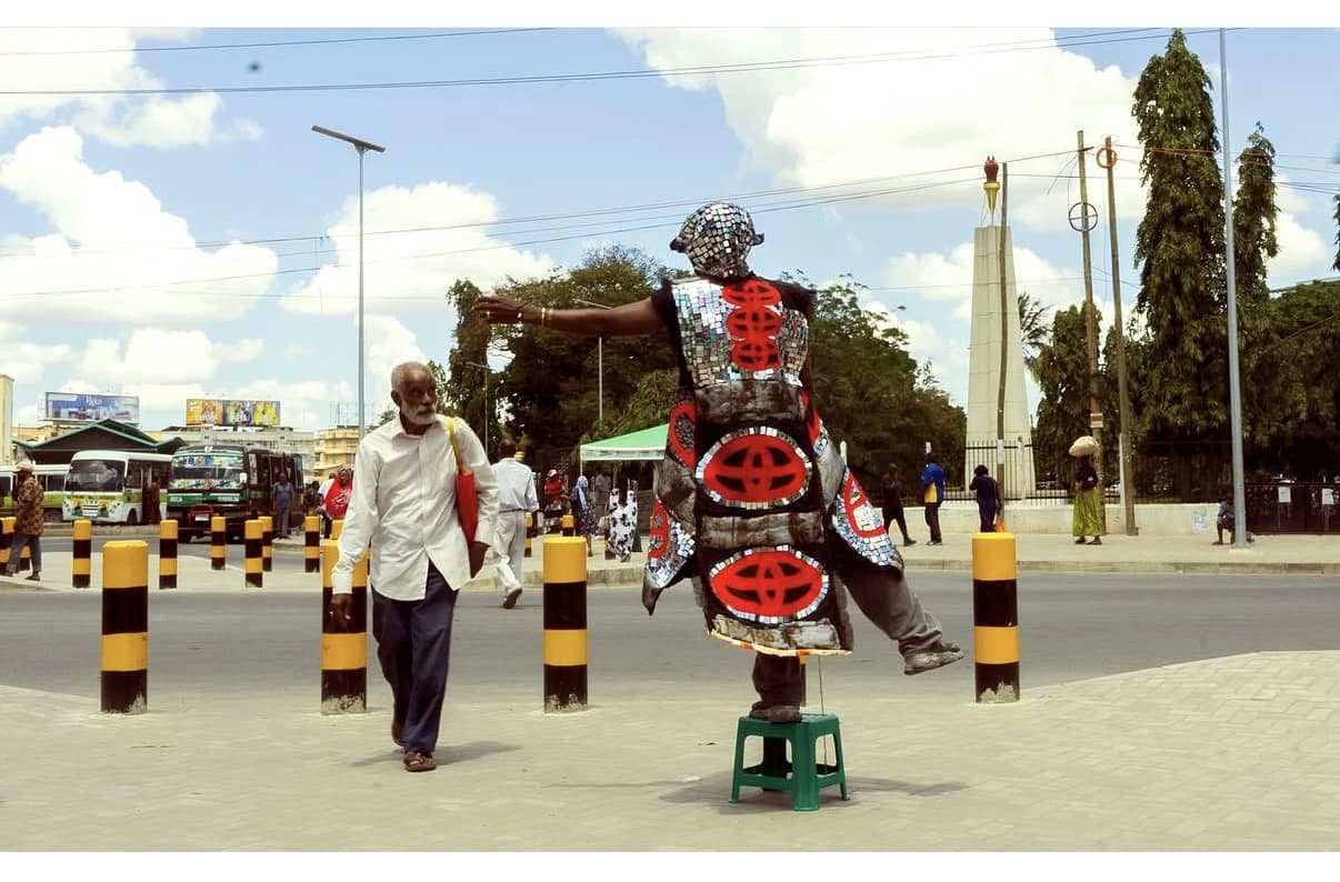 A costumed performer with circular red decorations walks through a city street